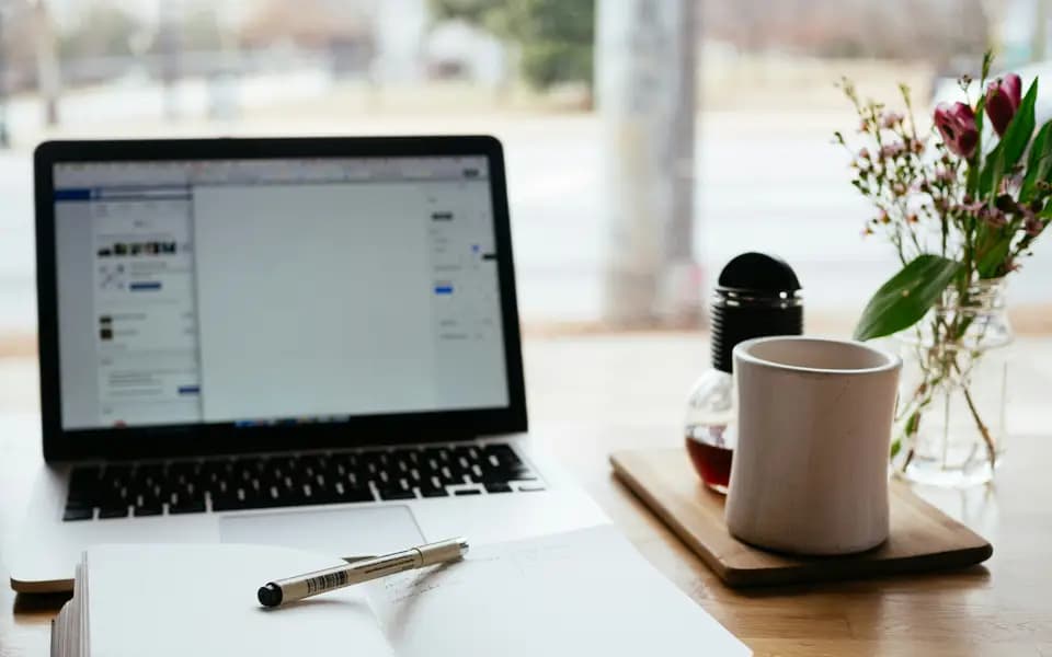 A laptop, notebook and mug on a study desk by a window